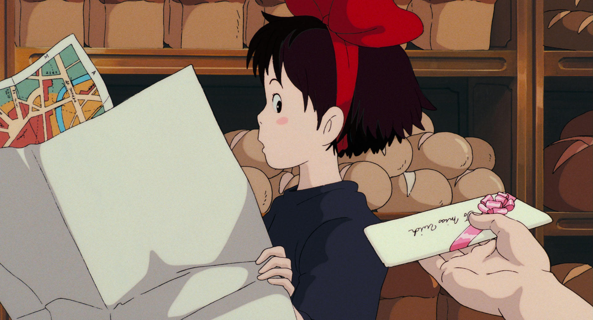 Kiki holds a large bag with a map sticking out while an unseen hand offers her a ribbon-tied envelope in a bakery